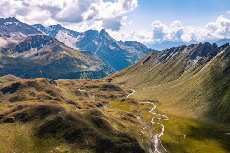 Vue aérienne en direction de la Capanna Motterascio, un chalet d'alpage situé sur le plateau de la Greina à Blenio, dans les Alpes suisses. Une crête rocheuse sur la droite conduit le regard vers la rivière qui coule sinueusement dans le paysage vallonné.
